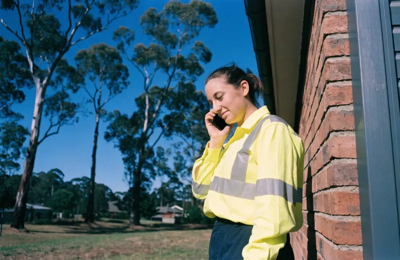 Australian tradesperson in hi-vis taking a phone call beside a brick wall
